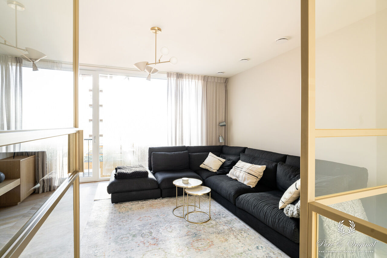 Interieur met een blauwe zetel in een wit interieur, gecombineerd met gouden accenten zoals deurframes en salontafel. Houten materialen zorgen voor warmte en balans in een elegante, lichte ruimte.