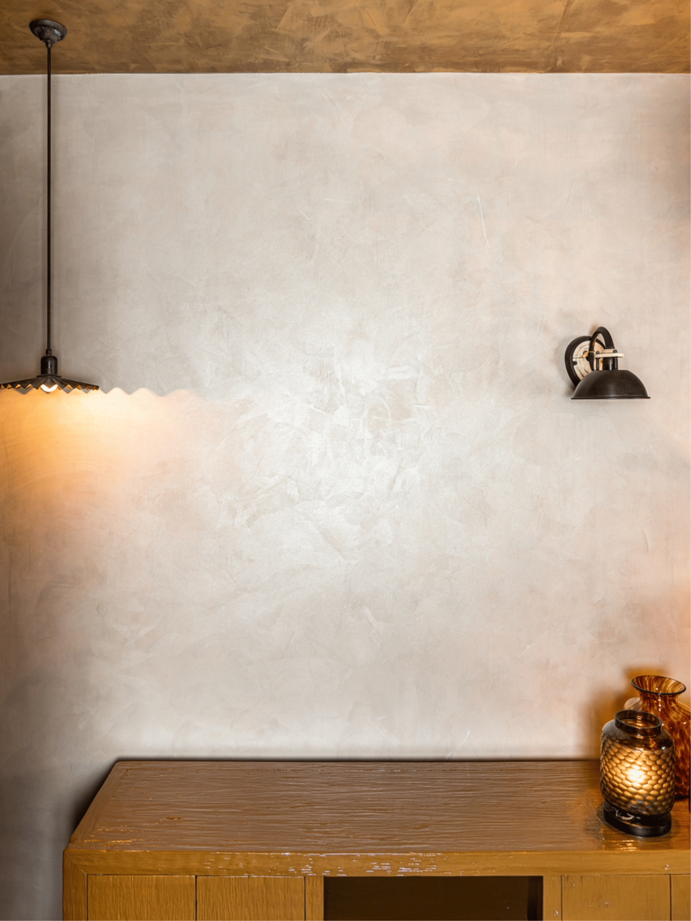 Interieur met lichte glinsterende verftechniek op muren en plafond in gebroken wit en goudtinten. Sfeervolle wandlampen, houten kastje en kaars zorgen voor een warme en elegante uitstraling.