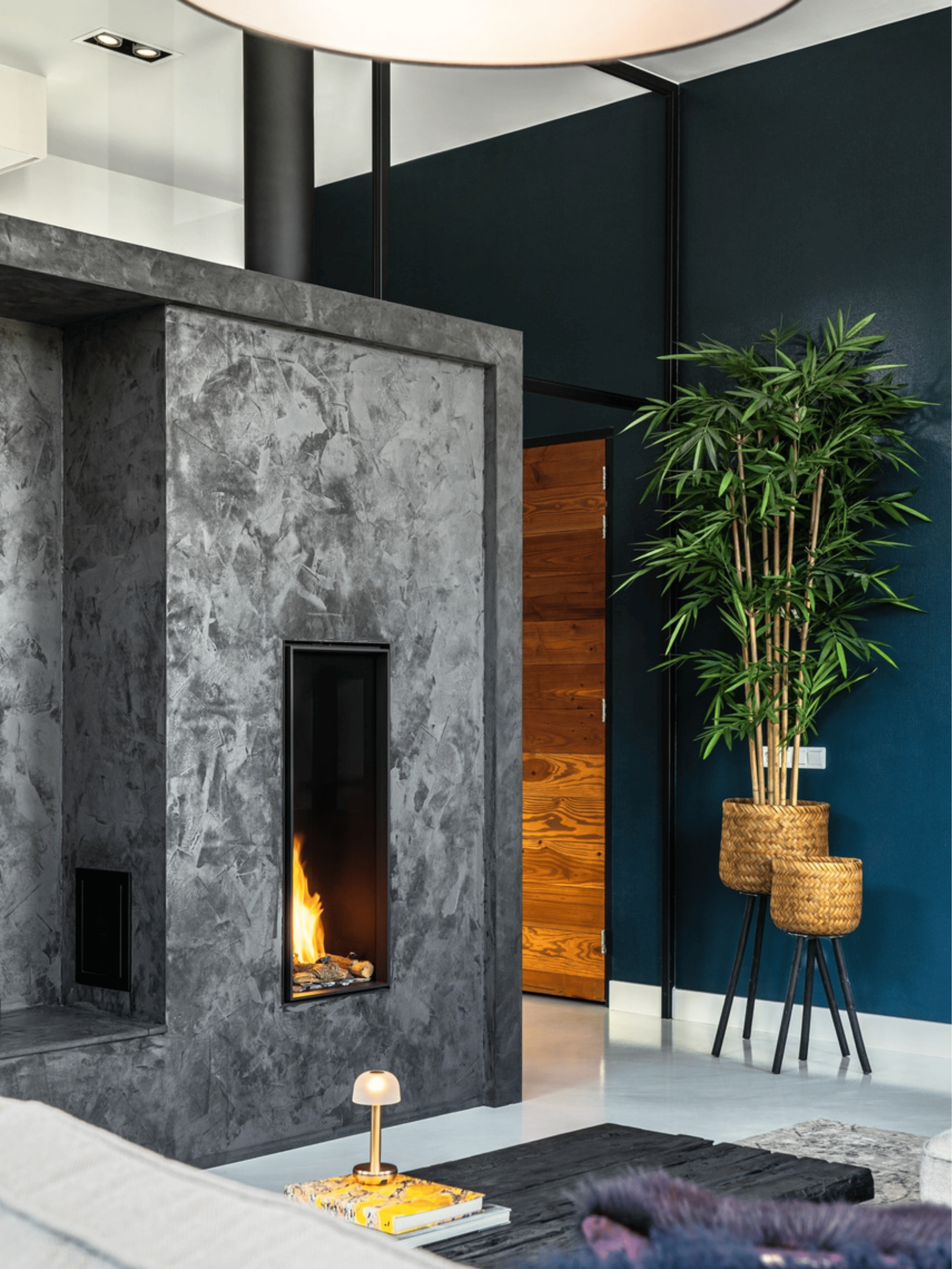 Moderne open haard met ruwe donkergrijze betonlook afwerking en luxueuze strakke vorm. Gecombineerd met donkerblauwe muren en veel groene planten voor een stijlvol industrieel interieurcontrast.