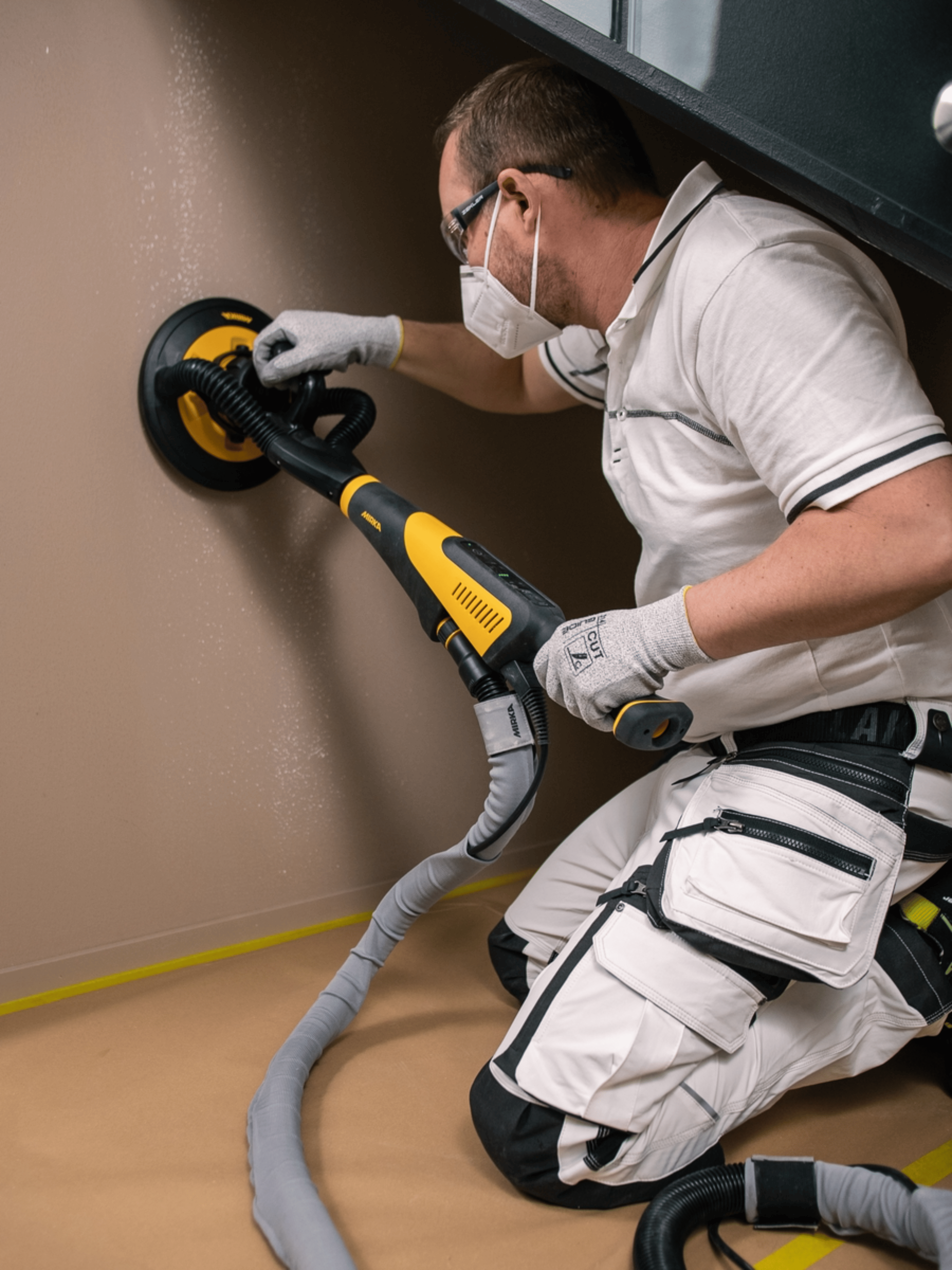 Vakman schuurt gebukt de muur onder een trap met een Mirka schuurmachine en stofafzuiging. Stofarm schuren voor een perfect voorbereide ondergrond voor schilderwerken en decoratieve wandafwerkingen.