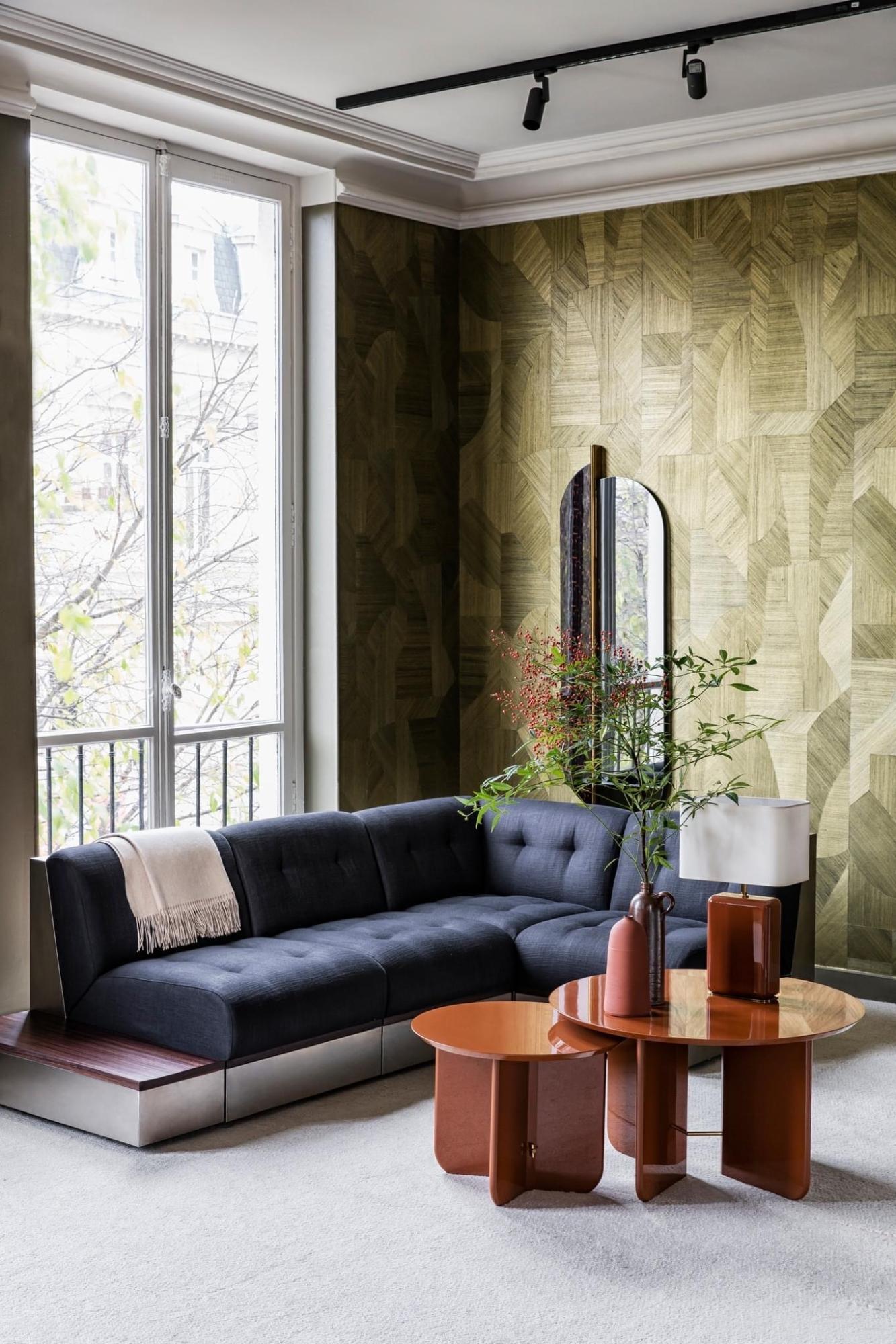 Showroom appartement RED met geometrisch groen geweven Arte Walls behang, blauwe zetel, donker roodbruin houten salontafel en planten. Modern interieur met warme materialen en natuurlijke accenten.