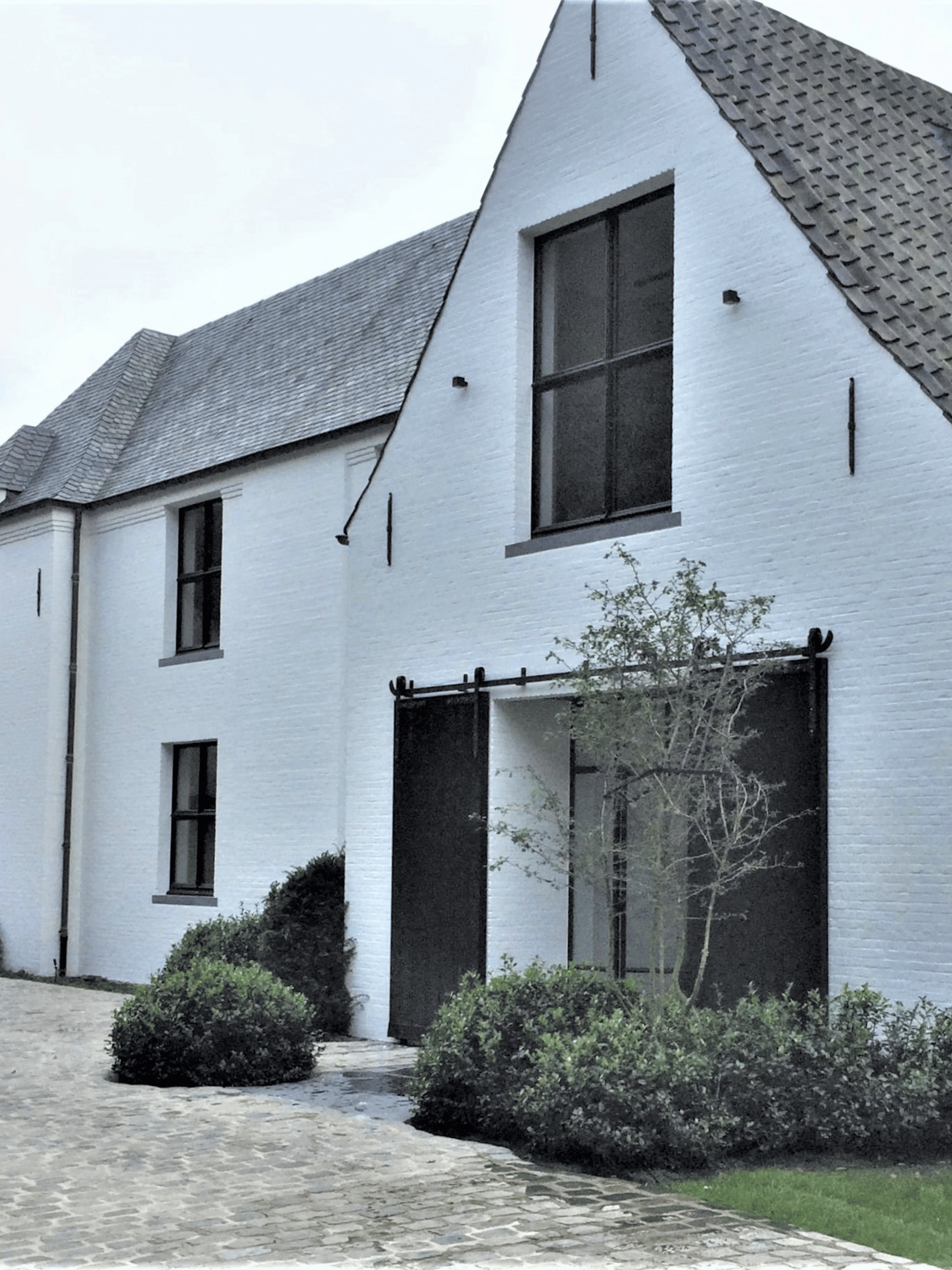 Moderne hoeve met een witte gekaleide gevel, grote zwarte aluminium raampartijen, een zwart dak en een groene tuin met weelderige beplanting. Duurzame kalei zorgt voor een strakke en tijdloze gevelafwerking.