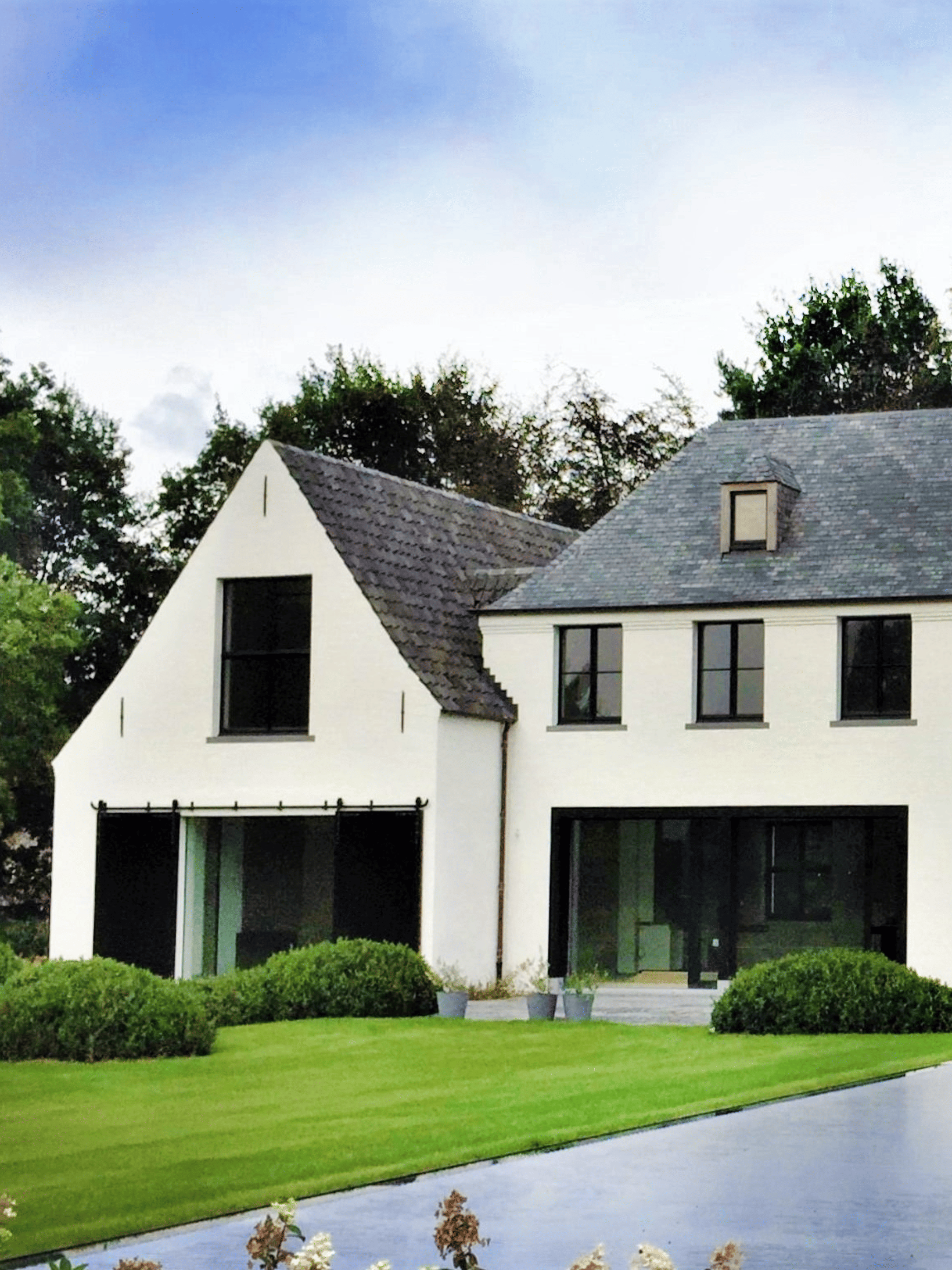 Moderne hoeve met een witte gekaleide gevel, grote zwarte aluminium raampartijen, een zwart dak en een groene tuin met weelderige beplanting. Duurzame kalei zorgt voor een strakke en tijdloze gevelafwerking.