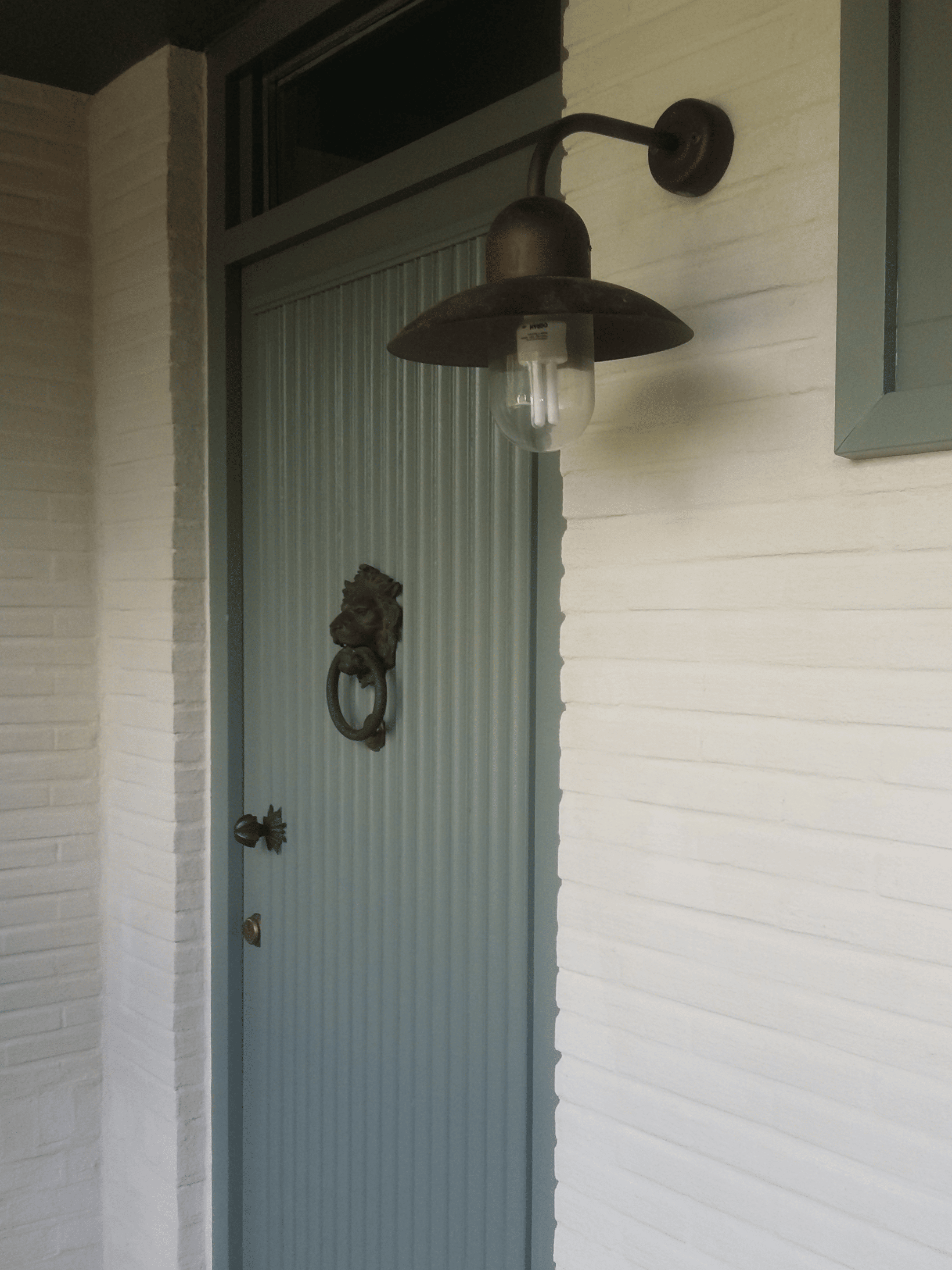 Close-up van een zachtgroene voordeur in een gekaleide gevel met bronzen wandverlichting. Landelijke gevelafwerking met duurzame kalei en stijlvolle entree.