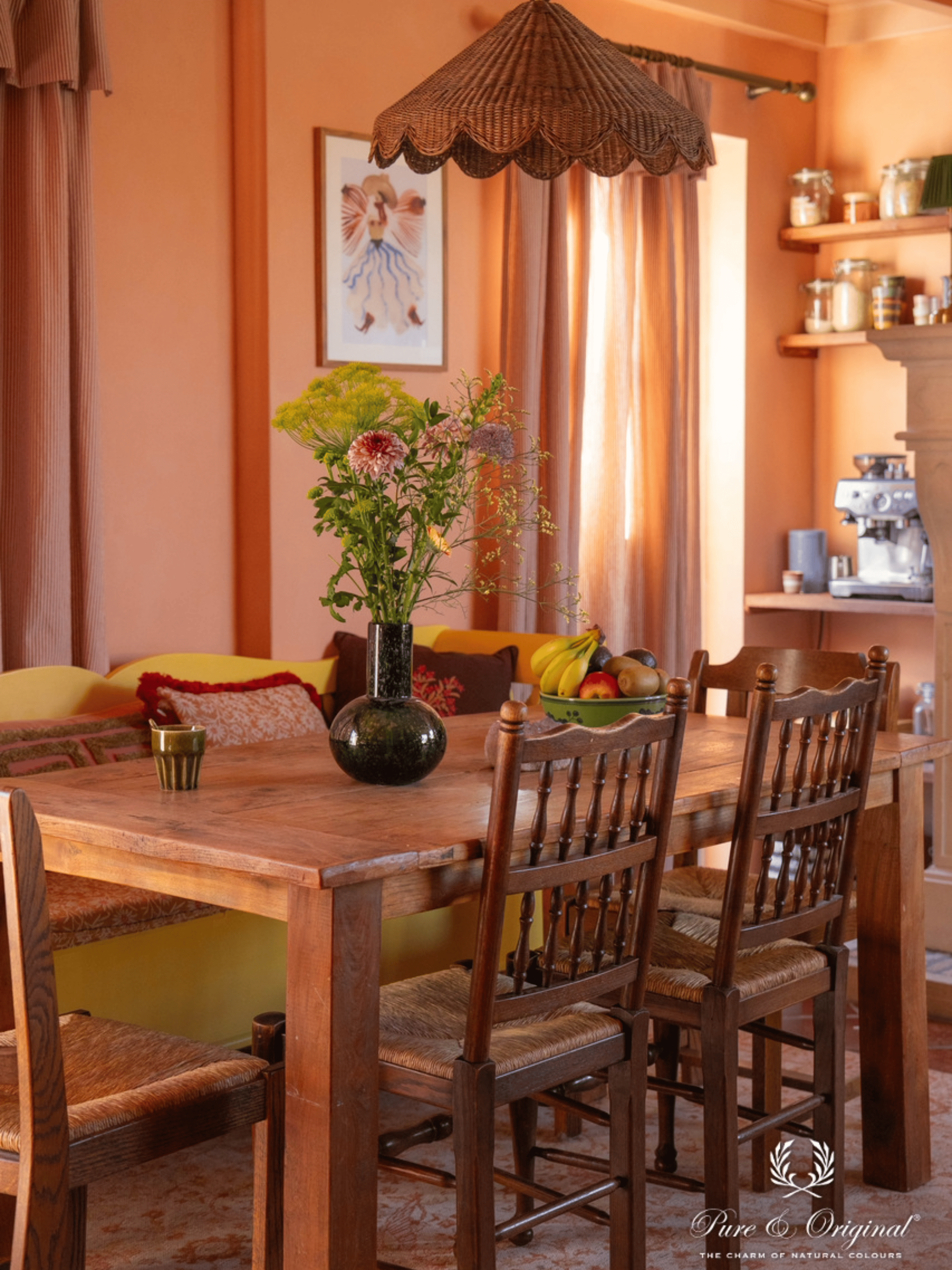 Eetkamer plaats met meerdere kleuren in het interieur. Een oranje zalm kleur zowel op de muren en het plafond. Een donkerdere tint hiervan als stoffering voor de gordijnen en een gele stoffen zetel als eyecatcher in het geheel. Donker bruin hout voor warmte en een rustig geheel.