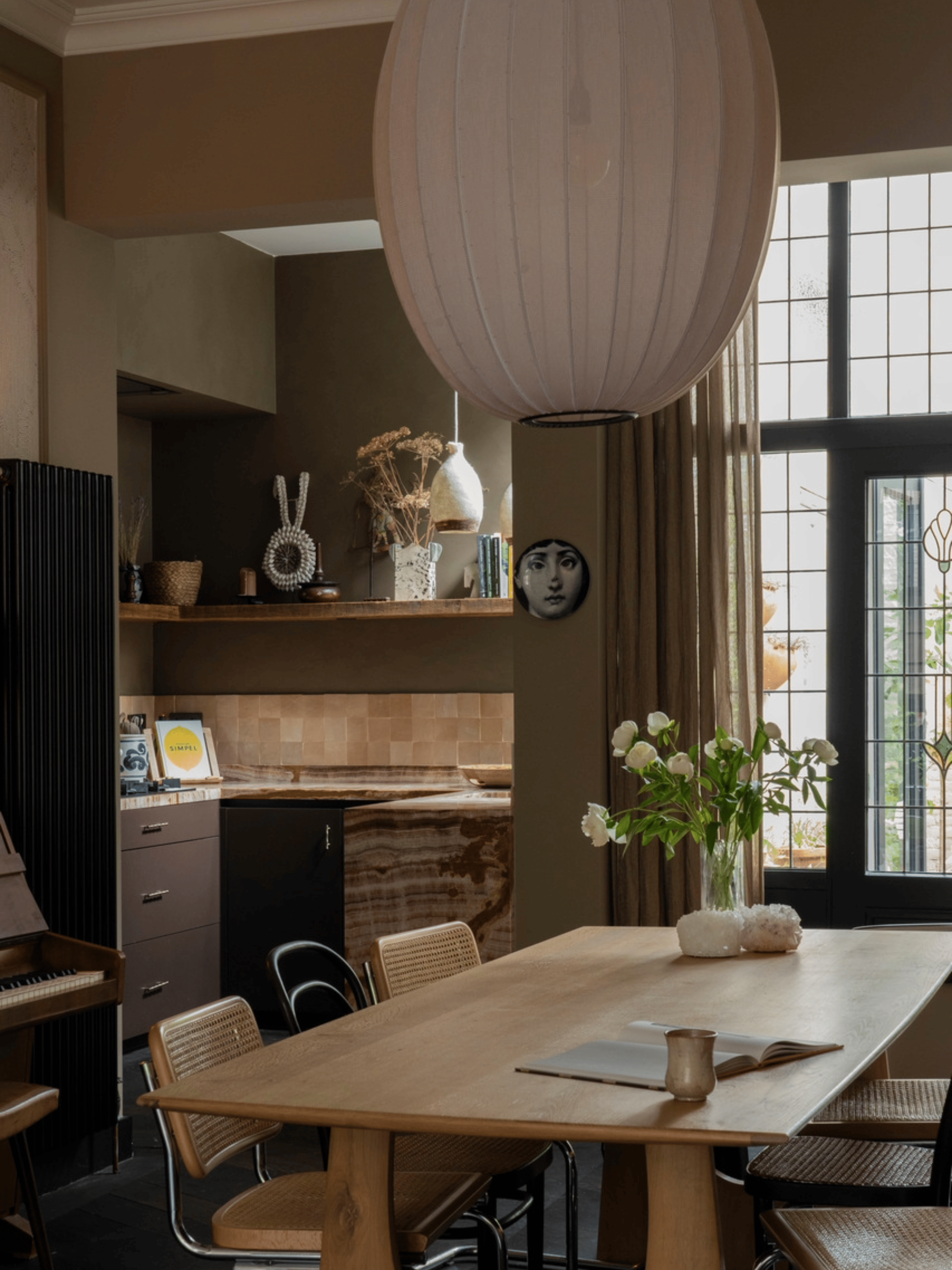 Landelijke eetkamer met bordeauxkleurige keukenkasten, zacht beige-groene wandafwerking en een houten eettafel. Witte rozen als decoratie zorgen voor een frisse en elegante, warme sfeer in het interieur.